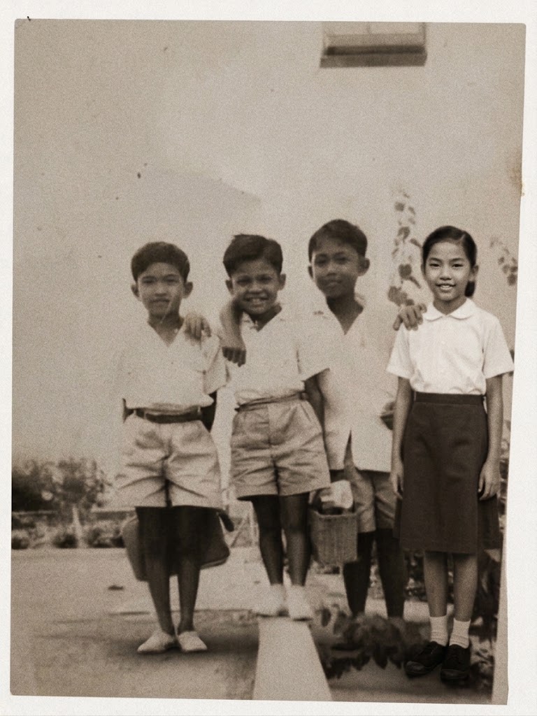 Four schoolchildren outside a building - three boys in white shirts and shorts, one carrying a basket, and a girl in a white blouse and dark skirt - a family photograph from an earlier Singapore